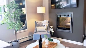 Un salon moderne avec une table basse ronde, un fauteuil gris, une plante en pot, une lampe sur pied et un téléviseur à écran plat fixé au mur.