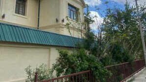 Un edificio de dos pisos con techo verde, cerca metálica roja y líneas eléctricas.