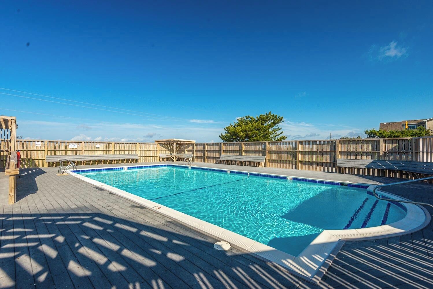A rectangular swimming pool with a wooden deck surrounding it, a wooden fence, and a tree in the background.