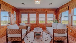 Una habitación con paredes de madera, ventanas amplias, mobiliario de mimbre y una mesa central.