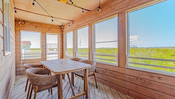 Terraza de madera con mesa y sillas, ventanas amplias con vista a vegetación e iluminación con luces colgantes.