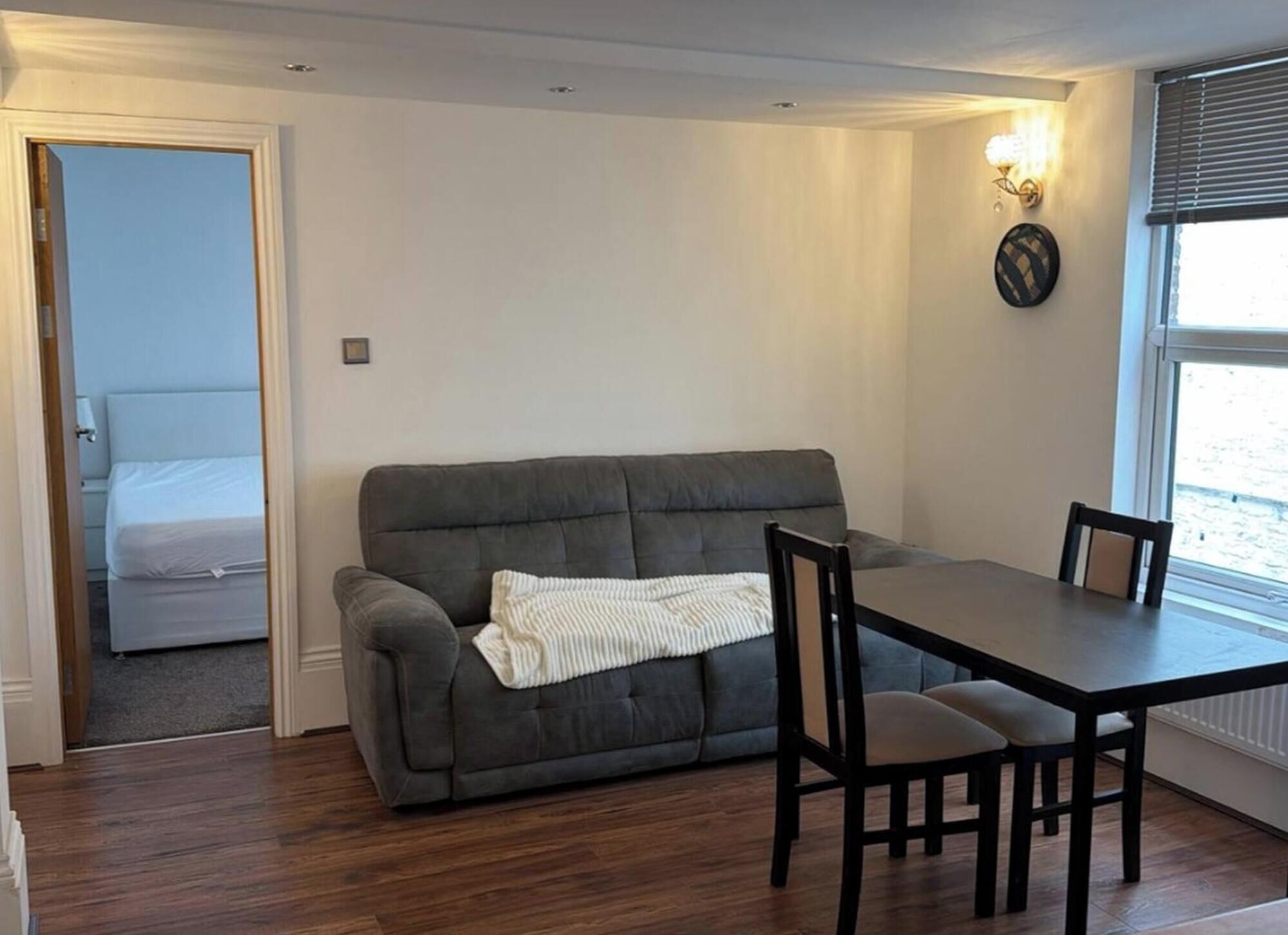 A room with a grey sofa, a dining table with chairs, a small window with blinds, and a door leading to another room.