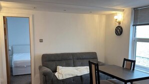 Ein Zimmer mit grauem Sofa, einem Esstisch mit Stühlen, einem kleinen Fenster mit Jalousien und einer Tür zu einem anderen Raum.