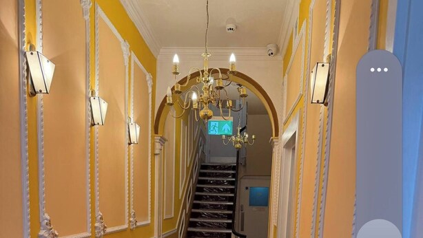 Ein Korridor mit Marmorboden, dekorativen Wandverzierungen, einer Chandelüster und einer Treppe, deren Ende eine blaue Tür versperrt.