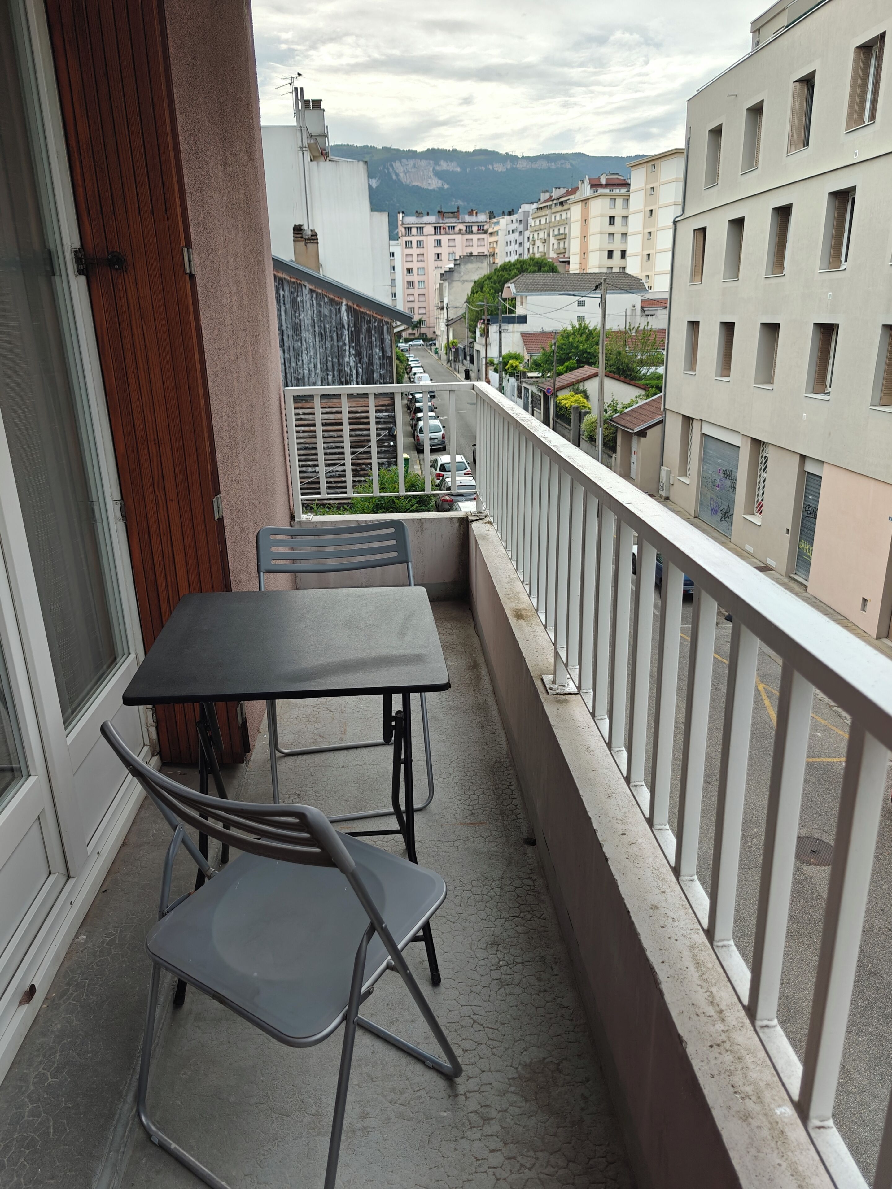 Un balcón con mesa y sillas, con vistas a una calle con coches aparcados y edificios.