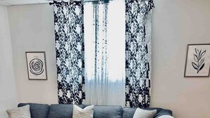 A living room with a grey sofa, a glass coffee table, a lamp on a side table, and patterned curtains. - Ship & Shore | Downtown Rooftop Deck | Fireplace (St Joseph)