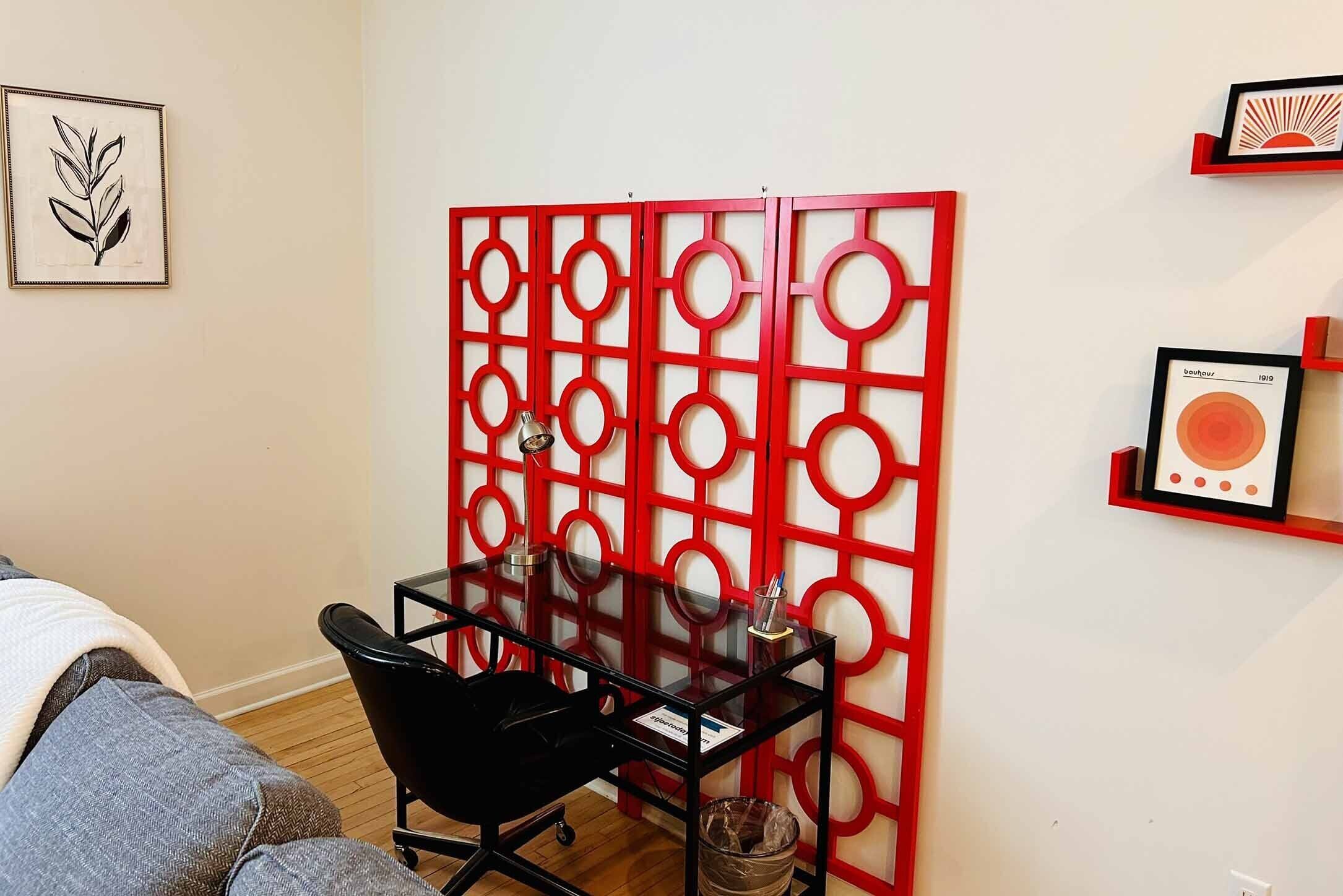 Una habitación con un escritorio de vidrio, una silla negra, un panel rojo y cuadros en las paredes.