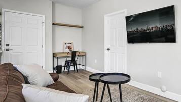Ein Wohnzimmer mit einem braunen Sofa, einem kleinen runden Tisch, einem Schreibtisch mit Stuhl und einem Stadtbild an der Wand.