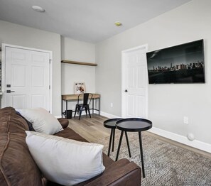 Ein Wohnzimmer mit einem braunen Sofa, einem kleinen runden Tisch, einem Schreibtisch mit Stuhl und einem Stadtbild an der Wand.