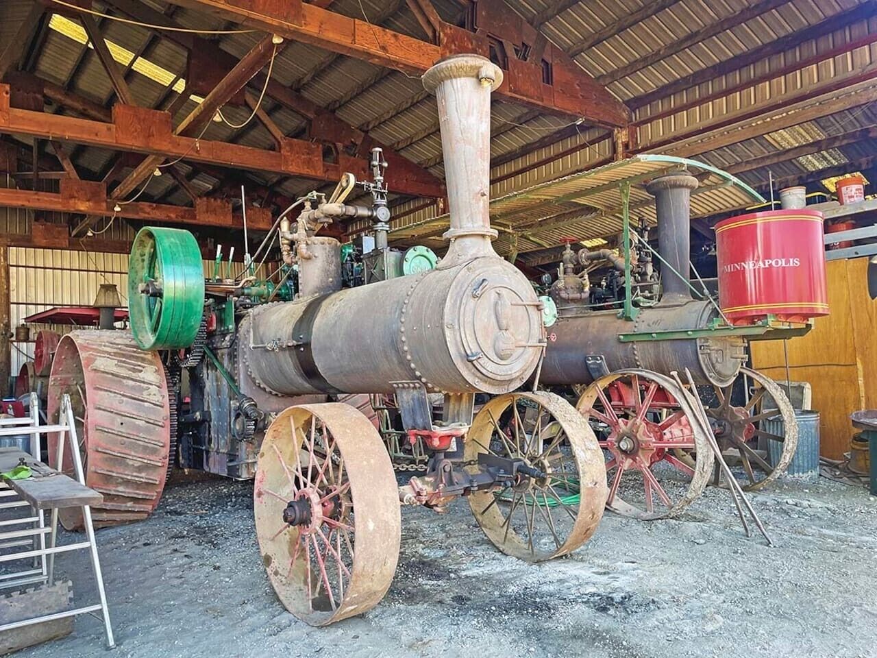 A steam locomotive with large wheels and a tall chimney, parked inside a building with wooden beams and a metal roof.