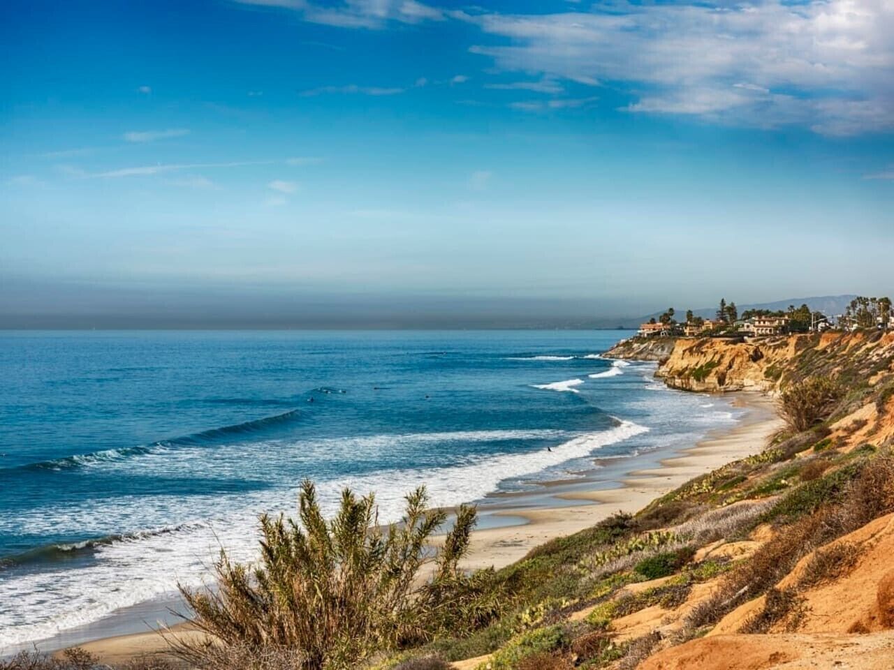 A coastal landscape with a beach, ocean waves, and a cliff with houses.