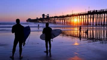 Silhouettes of people with surfboards on a beach at sunset, a pier in the background, and the ocean.