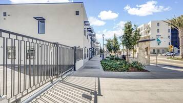 A modern building with a metal fence, a paved walkway, and a street with palm trees and other buildings in the background.