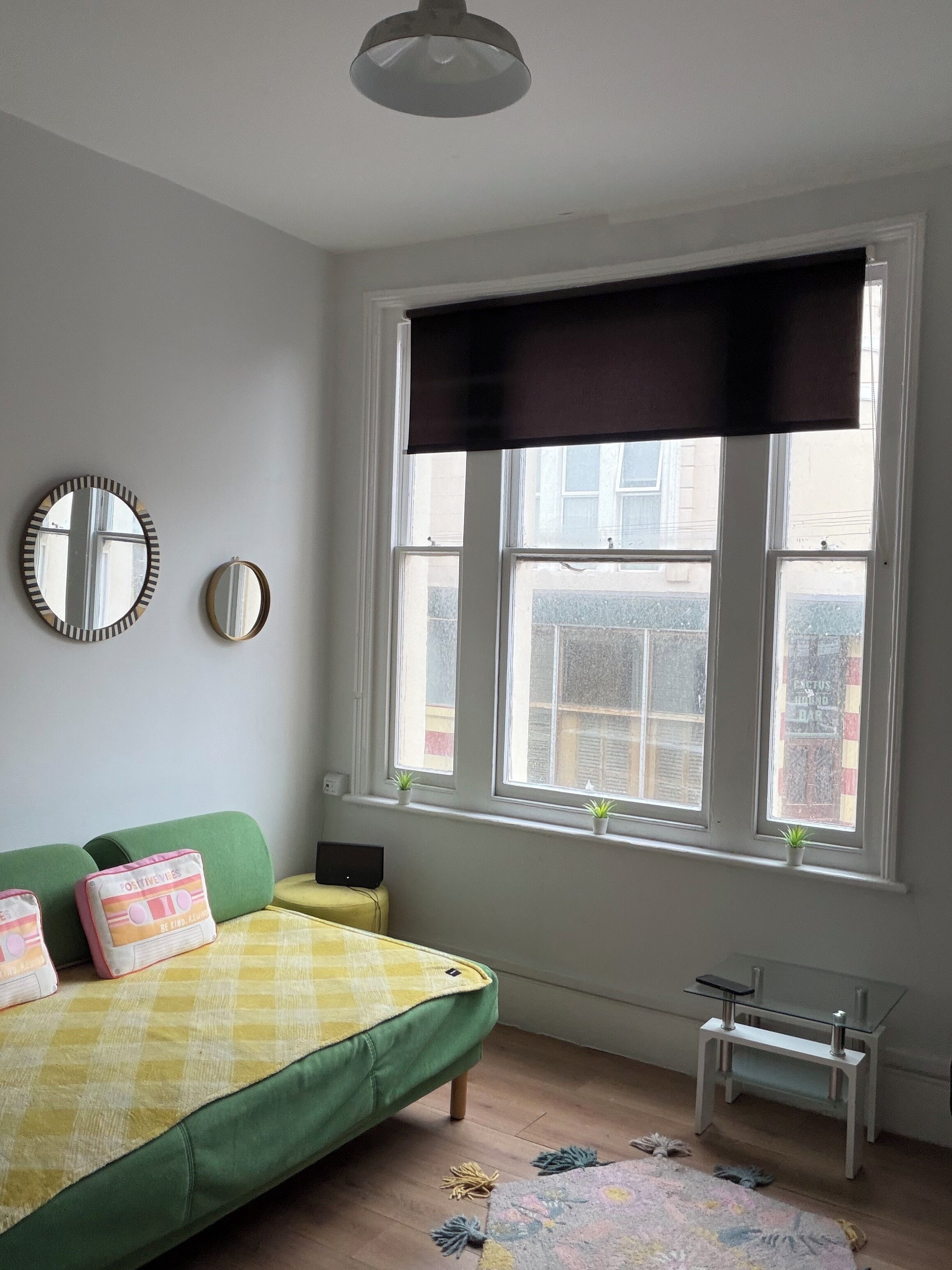 A room with a bed, two wall mirrors, a window with a blind, and a small table.