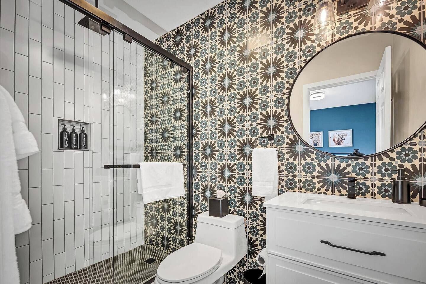 A bathroom with a shower, toilet, and sink, featuring patterned tiles and a round mirror.