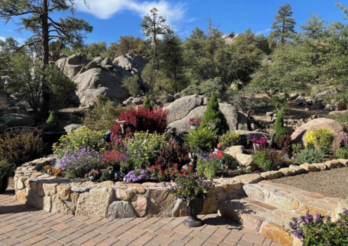 A landscaped garden with various plants and rocks, a paved walkway, and a backdrop of trees and hills.