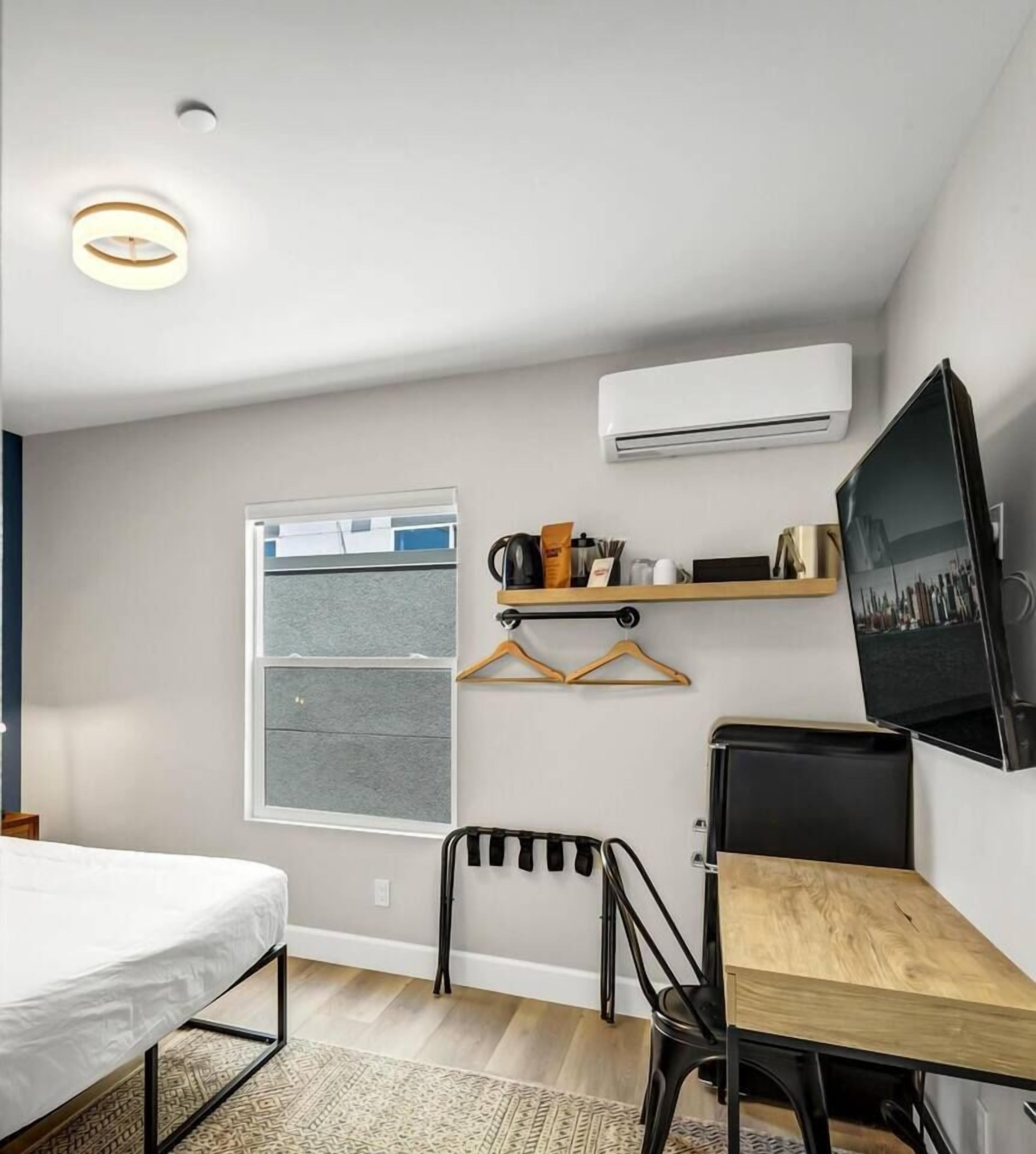A compact room with a bed, a small table, a chair, a TV, and an air conditioning unit.