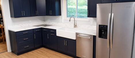 A modern kitchen with dark cabinets, a stainless steel refrigerator, a dishwasher, and a window with a view of greenery.