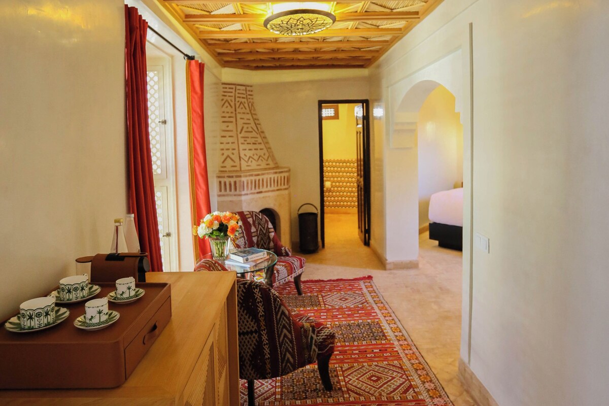 A room with a wooden ceiling, a dining area with a table and chairs, a red rug, and a bed in the background.