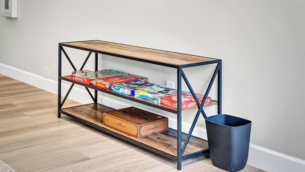 Ein hölzernes Konsolentisch mit Metallregalen, auf denen Brettspiele, ein Mülleimer und eine weiße Wand mit Kunstwerken stehen.