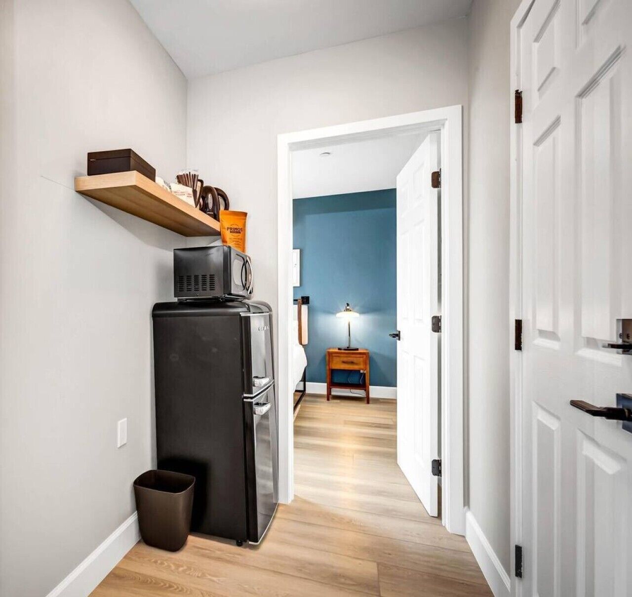 A hallway with a black refrigerator, a wooden shelf, and a trash bin.