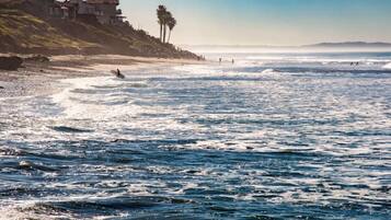 A coastal beach with waves, a clear sky, and a hillside with buildings.