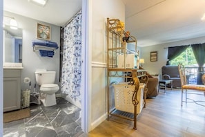 Un baño con inodoro, lavabo y cortina de ducha. En la zona de estar contigua hay una silla de madera y una mesa.