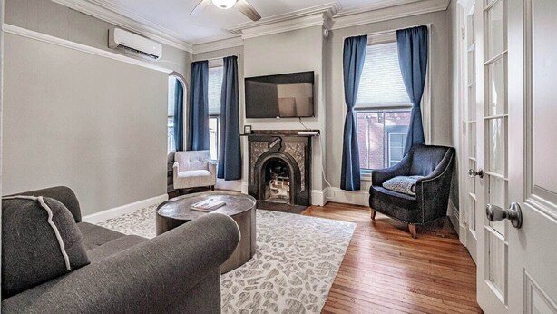 A living room with a fireplace, a ceiling fan, and a flat-screen TV. - Experience Portland’s charm in this beautifully restored 2-bed, 1.5-bath Victorian apartment! Located downtown, enjoy modern comforts, historic elegance, a private backyard, AC, and 1 free parking spot. Perfect for your Maine getaway! 🦞 (Portland)