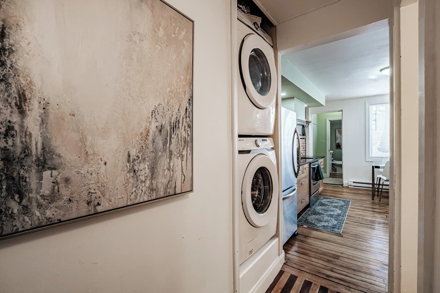A laundry room with a washing machine and dryer, a large abstract painting on the wall, and a wooden floor leading to a dining area.