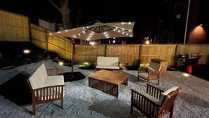 An outdoor patio area with wooden furniture, including chairs and a table, under a canopy with string lights. - Experience Portland’s charm in this beautifully restored 2-bed, 1.5-bath Victorian apartment! Located downtown, enjoy modern comforts, historic elegance, a private backyard, AC, and 1 free parking spot. Perfect for your Maine getaway! 🦞 (Portland)
