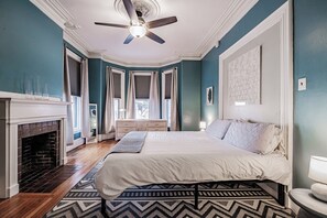 A bedroom with a bed, a fireplace, a ceiling fan, and a window with shutters.