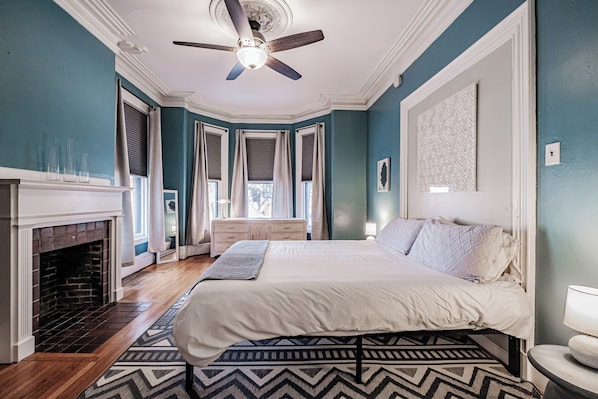 Un dormitorio con cama, chimenea, ventilador de techo y ventana con contraventanas.