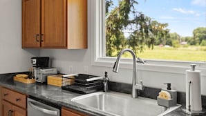 A kitchen with wooden cabinets, a stainless steel sink, a coffee maker, and a window with a view of greenery. - Charming Hudson Farmhouse with Fire Pit (Hudson)