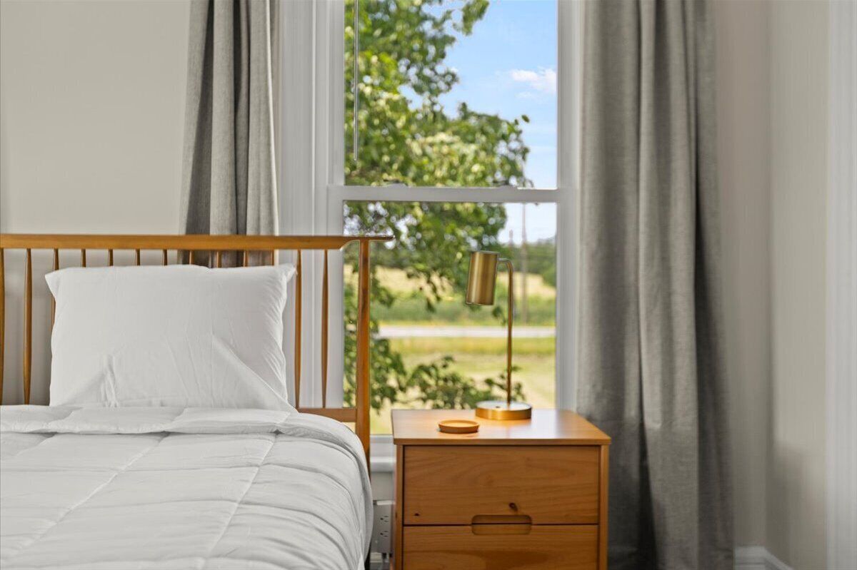 A bedroom with a bed, a nightstand, a lamp, and a view of greenery through the window.
