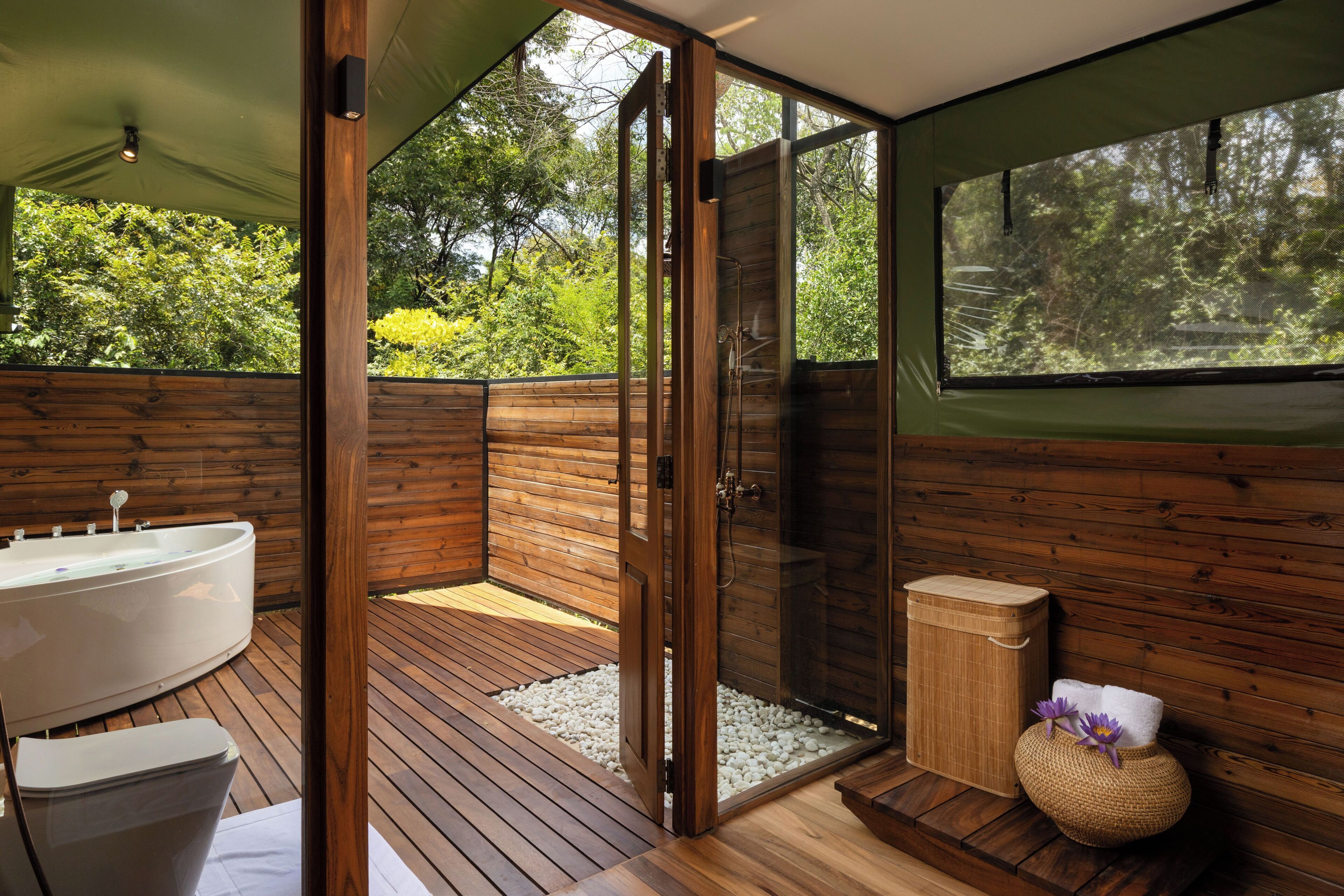 Un baño con piso de madera, una bañera, una ducha con mampara de vidrio y un ventanal con vistas a la vegetación.