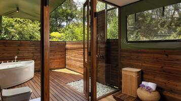 Un baño con piso de madera, una bañera, una ducha con mampara de vidrio y un ventanal con vistas a la vegetación.