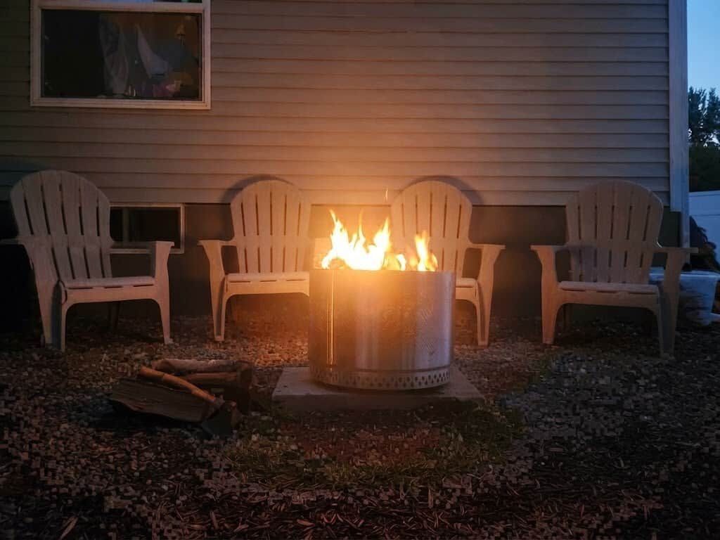 Ein Feuerkorb aus Metall mit Flammen, umgeben von weißen Kunststoffstühlen, auf einem Kiesplatz vor einem Haus.
