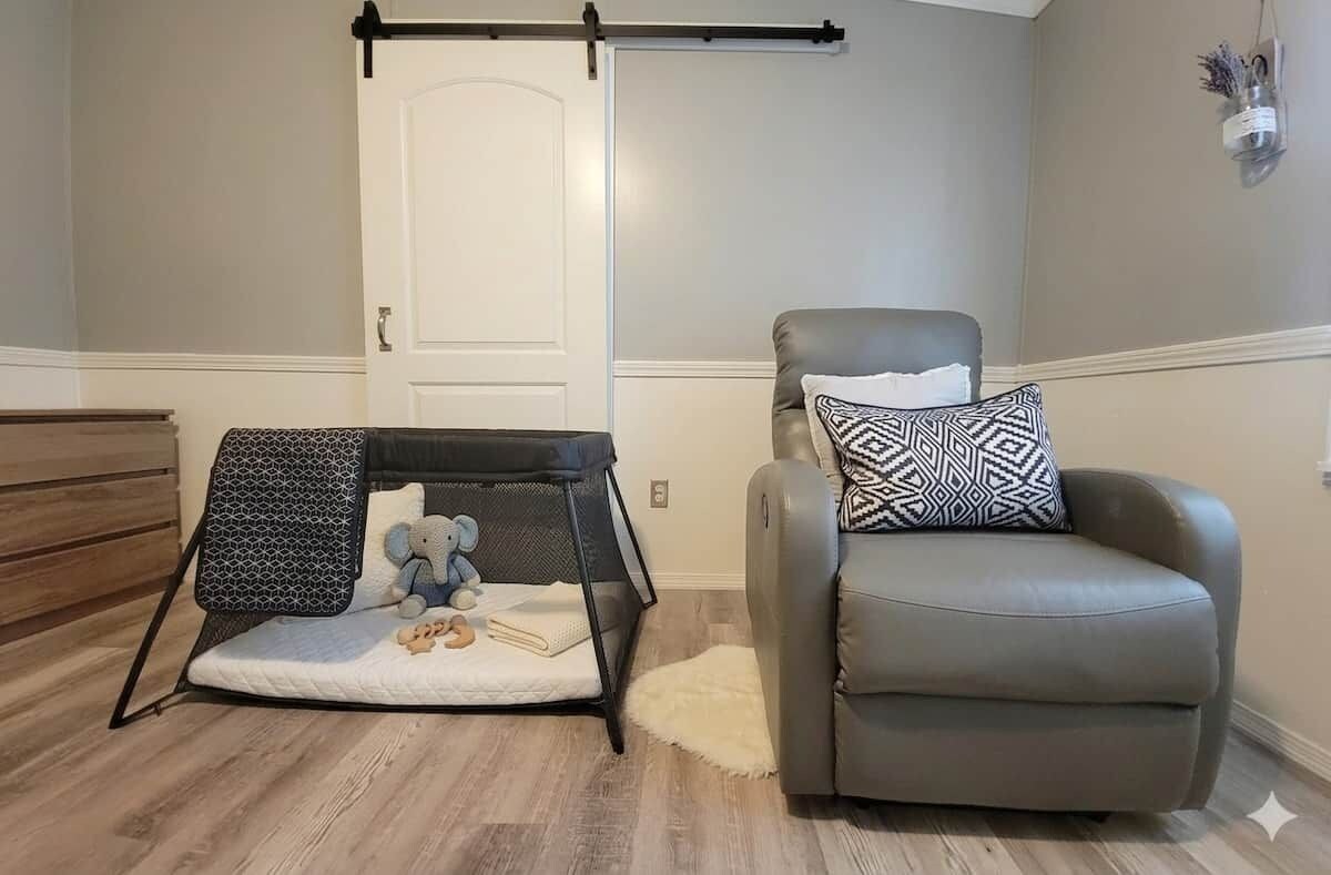Une pièce avec un parc pour bébé, un fauteuil gris et une porte blanche.
