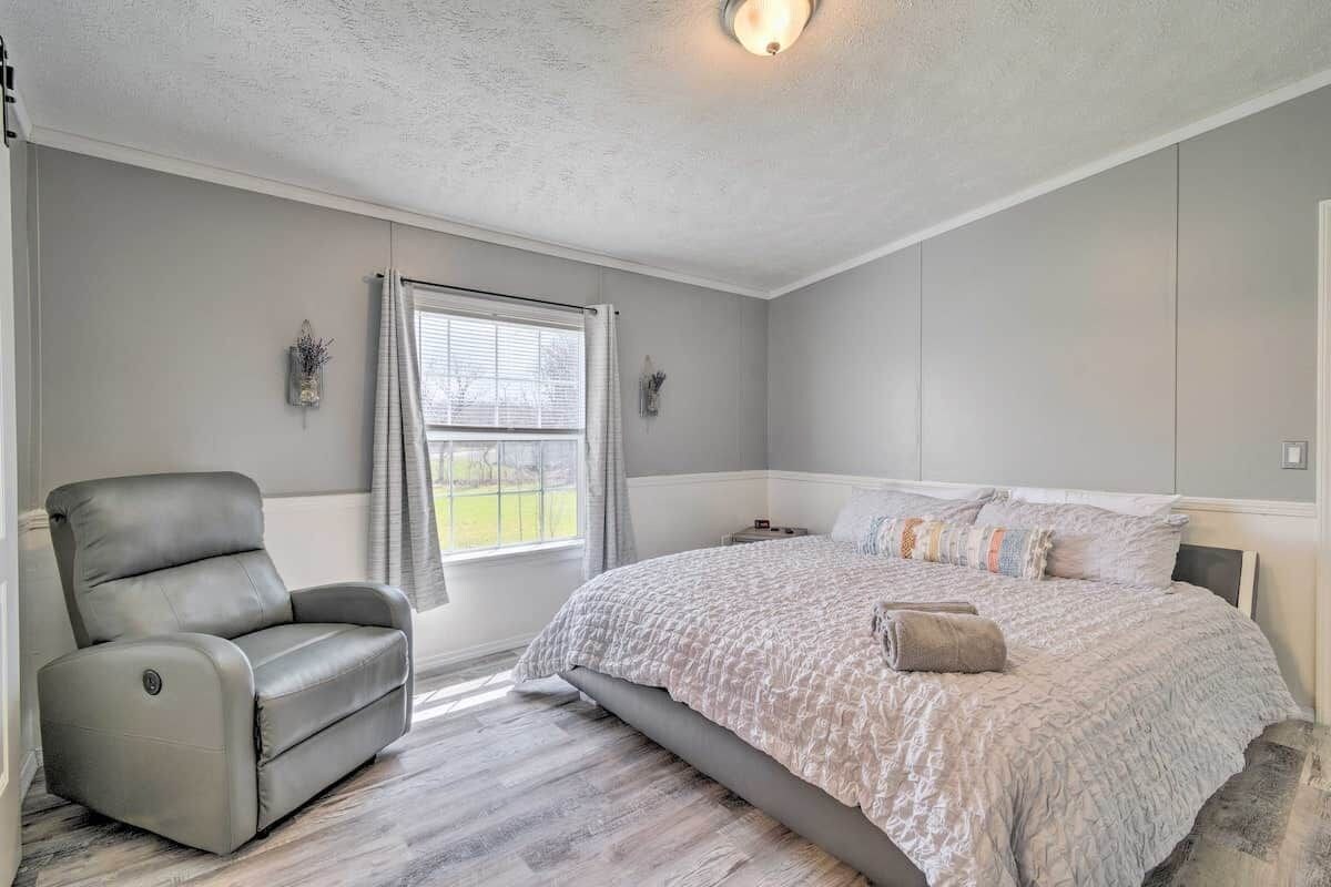 A bedroom with a bed, a reclining chair, a window with curtains, and a wall-mounted light fixture.