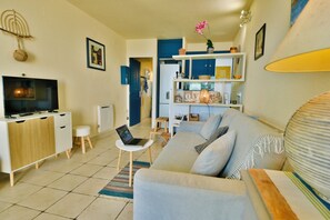 A living room with a sofa, a TV, a laptop on a small table, and a bookshelf. - Rarissime Studio with sea view & 17 m swimming pool | Absolute peace and quiet | 10 min from the beach (Hyères)