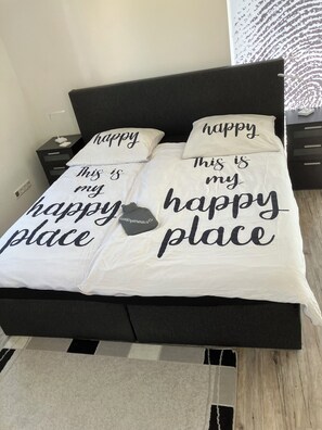 Ein Bett mit weißer Decke und schwarzen Kissen, auf denen jeweils der Schriftzug 'happy place' abgebildet ist.