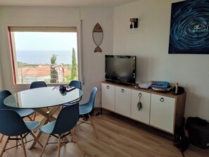 A modern room with a round dining table, blue chairs, a flat-screen TV, a cabinet with drawers, and a large window offering a view of the sea and buildings. - Eugenie's lighthouse (Llançà)