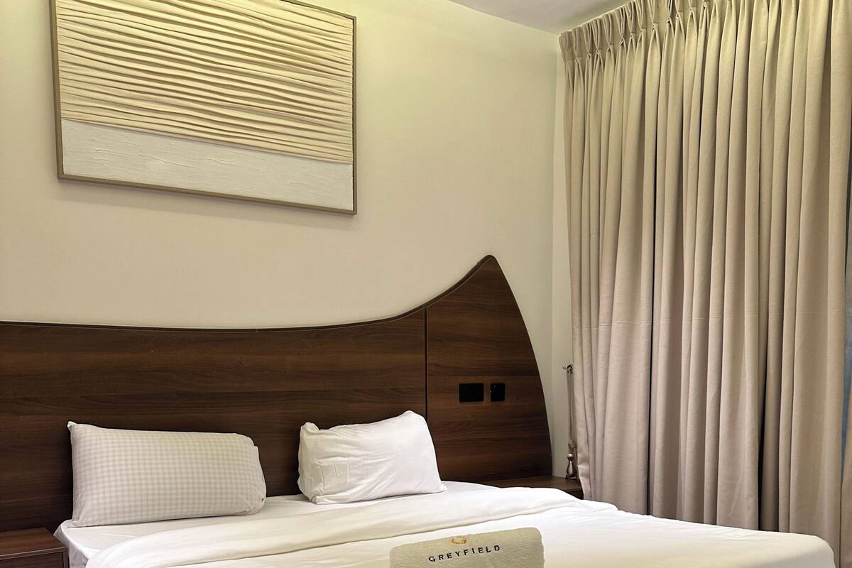 A neatly made bed with a headboard, a pillow, and a rolled towel with 'GREYFIELD' on it.