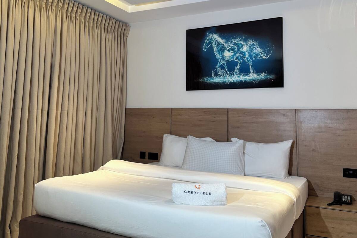 A modern hotel room with a large bed, a headboard, a wall-mounted artwork, and a bedside table.
