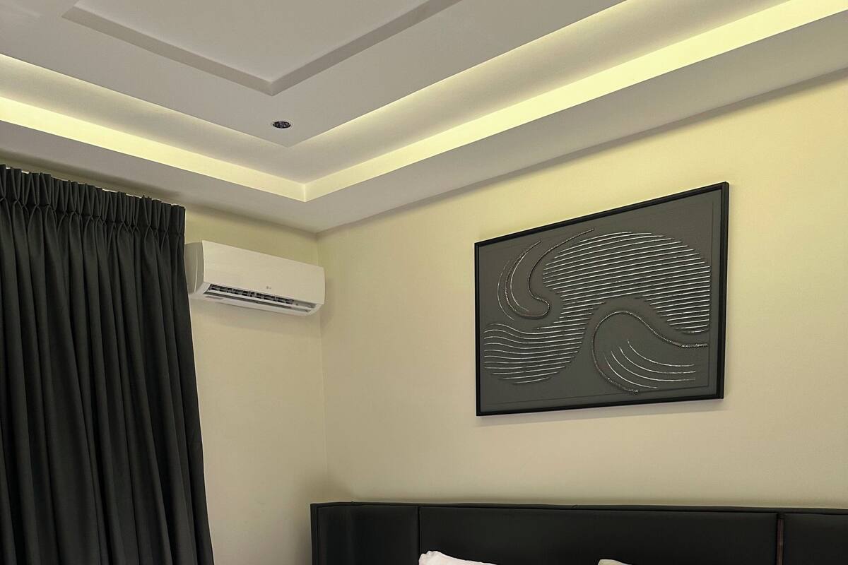 A modern hotel room with a large bed, a headboard, a wall-mounted air conditioner, and a framed artwork.