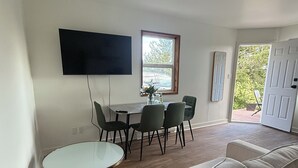 Een moderne woonkamer met een bank, een eettafel met stoelen, een flatscreen televisie aan de muur en een raam met uitzicht op groen.