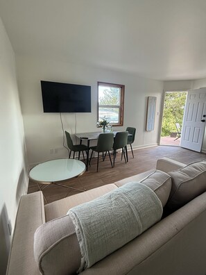 A modern living room with a sofa, a dining table with chairs, a flat-screen TV mounted on the wall, and a window with a view of greenery.