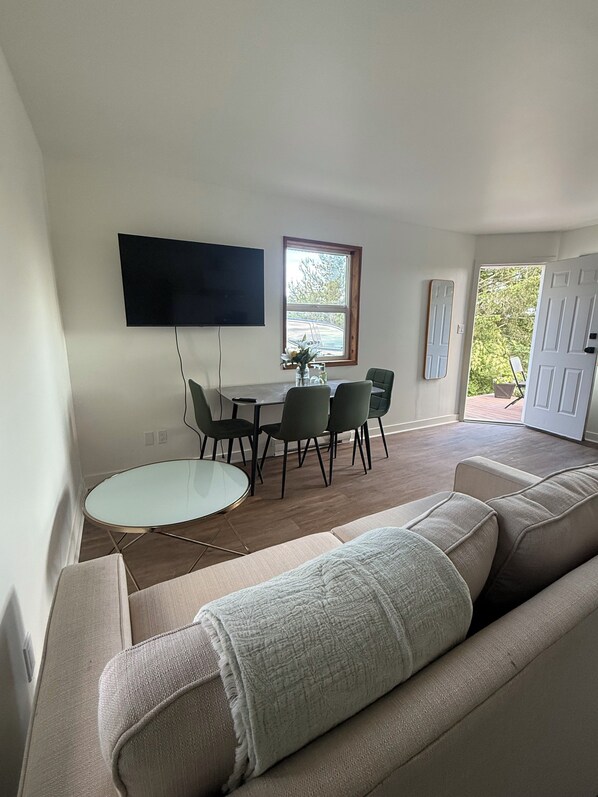 A modern living room with a sofa, a dining table with chairs, a flat-screen TV mounted on the wall, and a window with a view of greenery. - Beautiful View, Pet Friendly, Rural, Luna Blanca Lookout  (Sooke)
