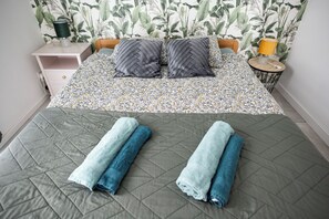 A neatly made bed with a patterned bedspread and pillows, a bedside table with a lamp, and a wall with a leafy wallpaper design. - Abracadabra Flat France Suisse near airport (Saint-Louis)