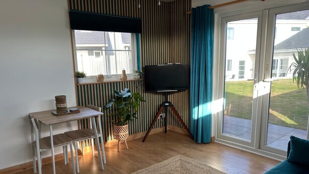 Olohuoneessa on jalustalla oleva televisio, ruokapöytä tuoleineen, sohva, suuri ikkuna, jossa on verhot, ja näkymä ulos.